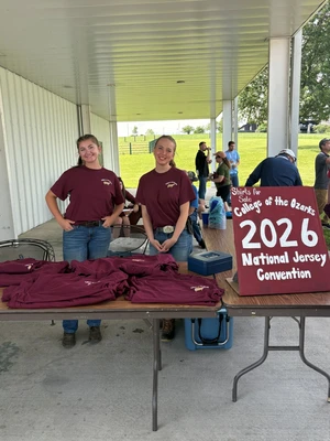 This photo depicts two girls standing behind a table selling red t-shirts. There is a sign beside them, also in red, with white letters that say "College of the Ozarks 2026 national Jersey Convention."